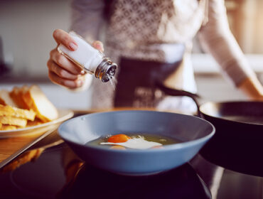 close up of caucasian woman adding salt in sunny side up eggs while standing in kitchen next to stove.