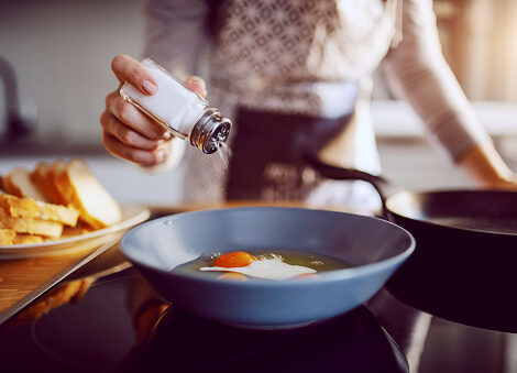 close up of caucasian woman adding salt in sunny side up eggs while standing in kitchen next to stove.