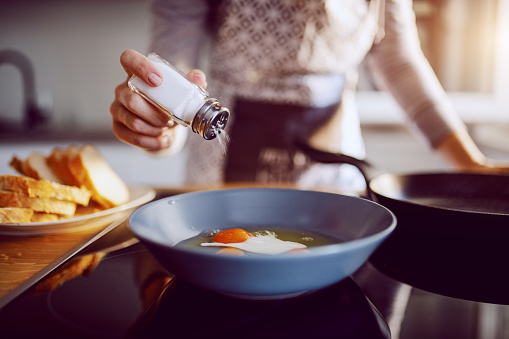 close up of caucasian woman adding salt in sunny side up eggs while standing in kitchen next to stove.